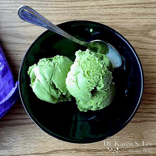 green tea ice cream in a black bowl on a wooden board with a spoon resting on it. 