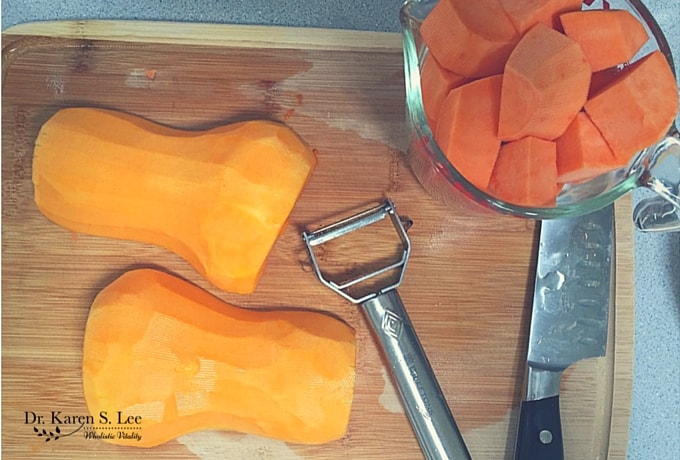 Peeled and split in half butternut squash on cutting board next to peeler, knife, and cubed butternut squash in bowl