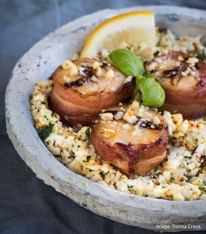 3 bacon wrapped scallops on a bed of white cauli couscous with a lemon wedge and basil on a greyish plate