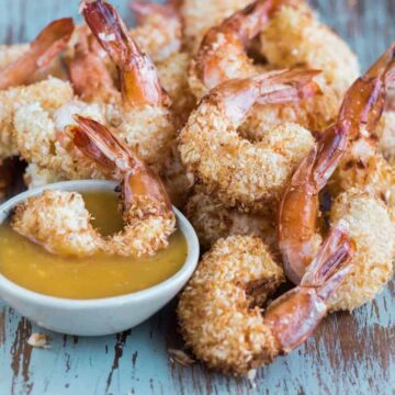crispy coconut shrimp on blue board with one shrimp dipped in yellowish sauce in a white bowl