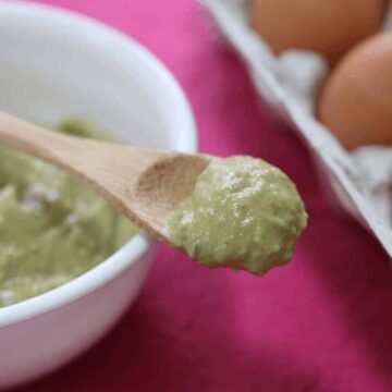 Avocado face mask on a wooden spoon over a whilte bowl on a pink placemat and eggs in the background