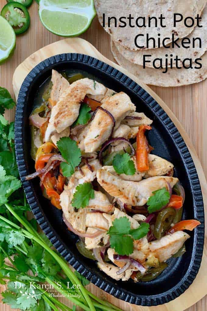 Cooked chicken strips and veggies on an oval black cast iron skillet on a wooden trivet surrounded by cilantro, lime and jalapeno peppers