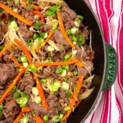 Brown beef slices with slivers of carrots and chopped scallions on a cast iron skillet with red and white stripe napkin in the background