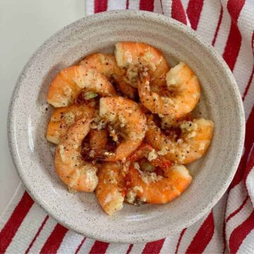 cooked shrimp in a white bowl, shells in a stainless bowl, on red and white striped kitchen towel