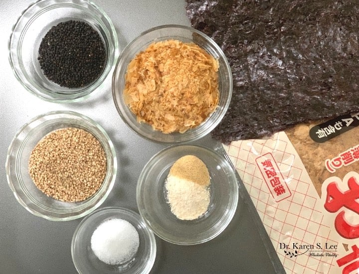 bowls of sesame seeds, bonito flakes, garlic and onion powder, salt, nori and bonito packages on a white table. 