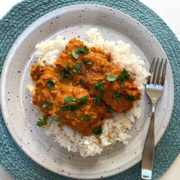 Salmon Tikka Masala in curry sauce over white rice on a speckled whitish plate on a round moss green placemat with a stainless steel fork on the right