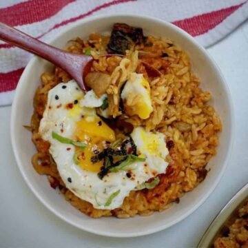 spoonful of kimchi pork fried rice with sunny side up egg on top above a white bowl filled the same on the table with red and white kitchen towel on it