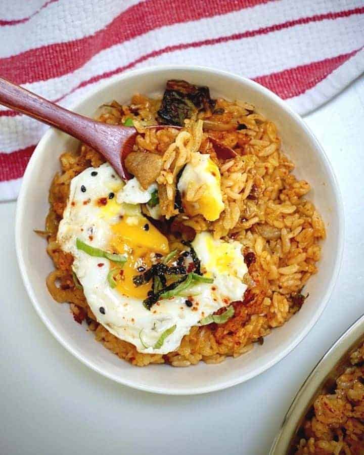 spoonful of kimchi pork fried rice with sunny side up egg on top above a white bowl filled the same on the table with red and white kitchen towel on it