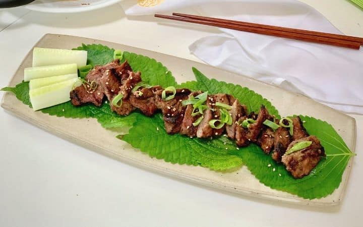 barbecued beef pieces on green vegetables on a cream color oblong shaped platter with brown chopsticks in the background