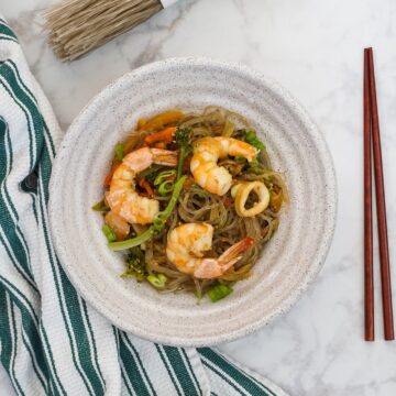 Light beige bowl with glass noodles with seafood and vegetables next to green white striped towel with brown chopsticks on right side