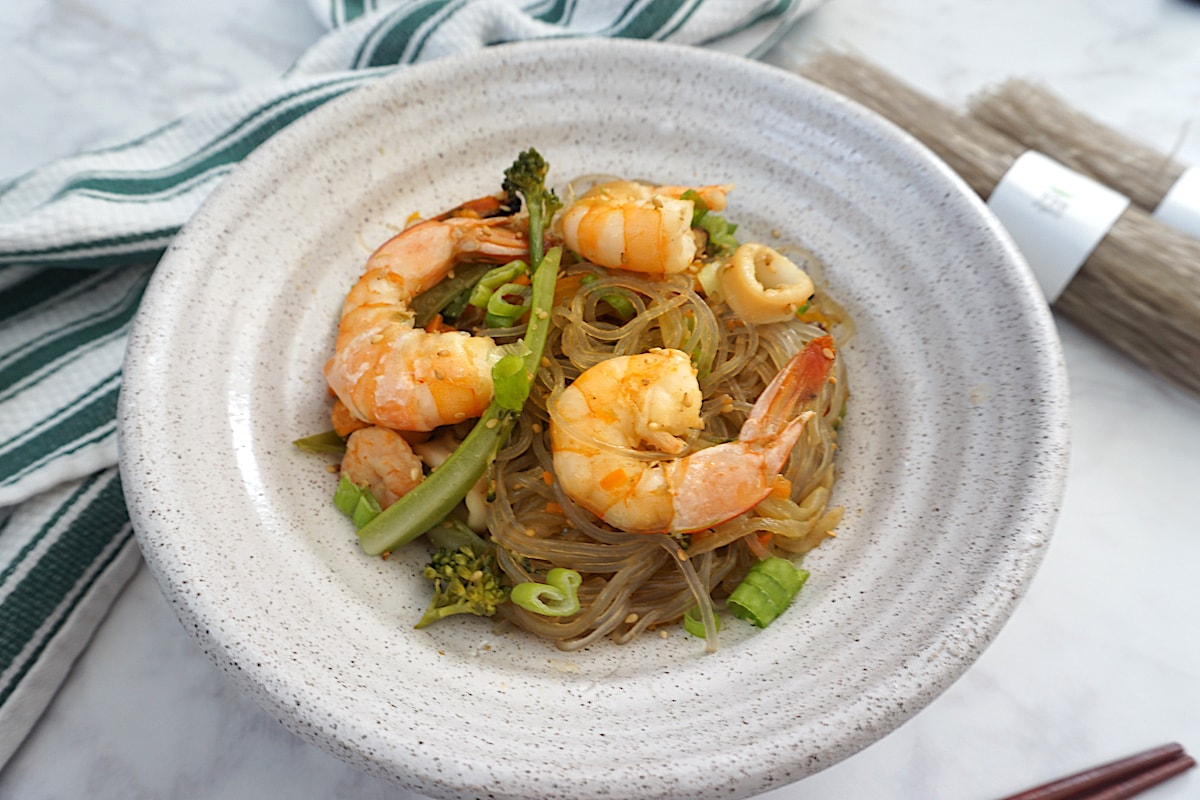 Light beige bowl with glass noodles with seafood and vegetables next to green white striped towel with brown chopsticks on right side