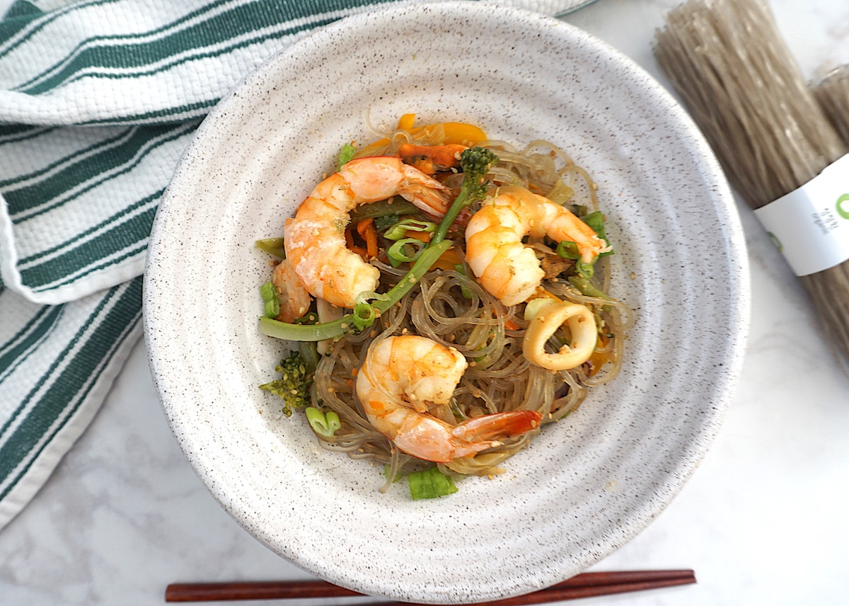 Light beige bowl with glass noodles with seafood and vegetables next to green white striped towel with brown chopsticks on right side