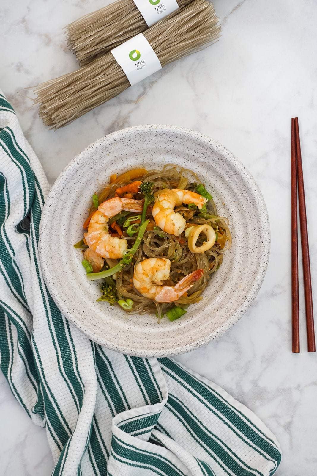 Light beige bowl with glass noodles with seafood and vegetables next to green white striped towel with brown chopsticks on right side