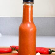 red hot sauce in glass bottle with black cap surrounded by red chili peppers and garlic on wood cutting board
