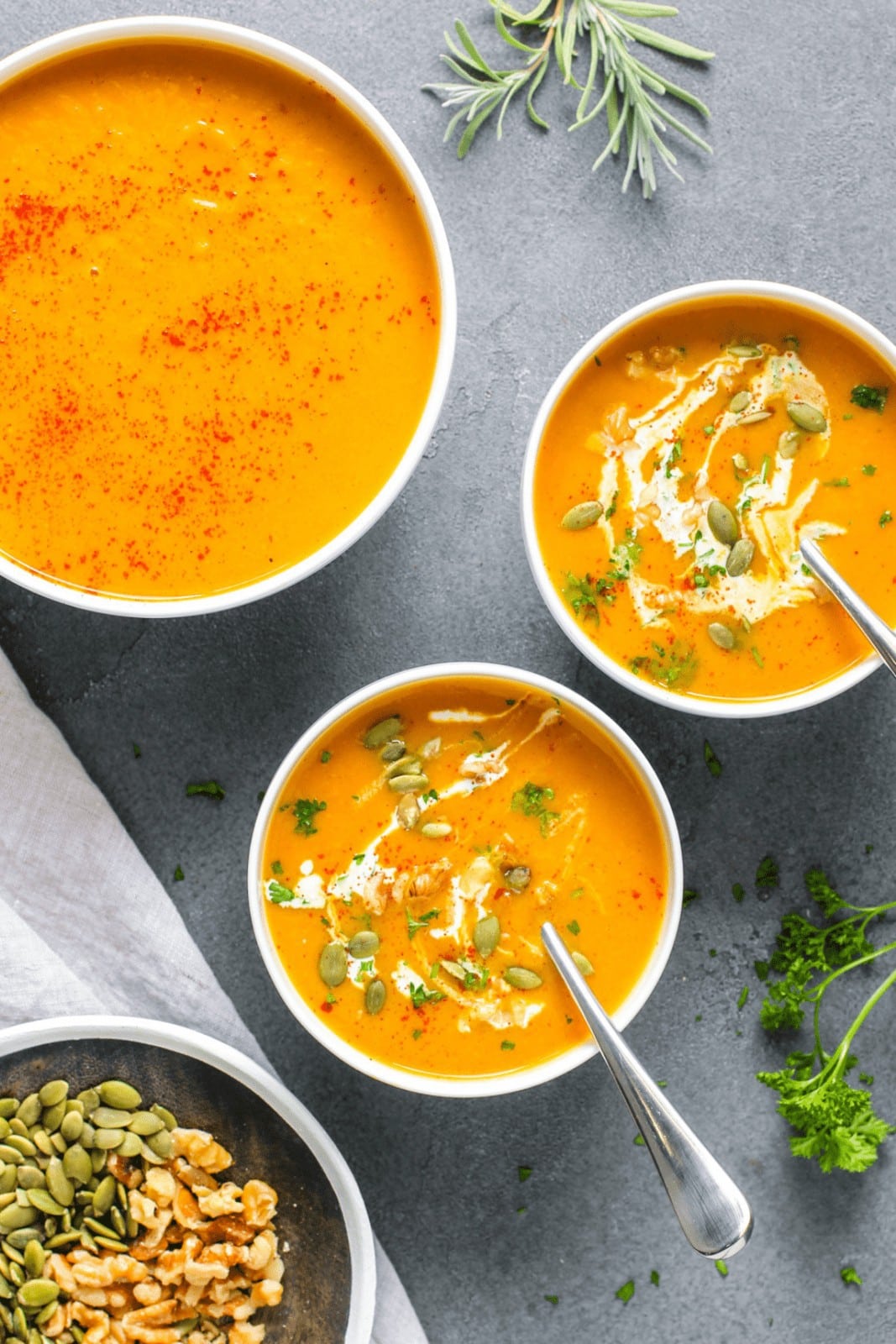 3 bowls of orange colored squash soup with cream and green garnish on grey background