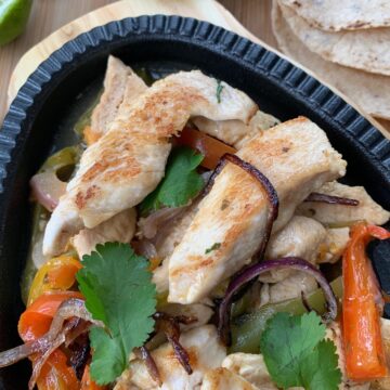 Chicken and vegetables plated on oval shaped black cast iron plate on wooden trivet surrounded by lime wedges and cilantro